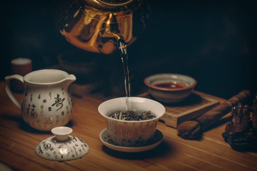 A beautiful copper kettle pouring hot water into a lovely china cup that has Rooibos Tea leaves waiting to be brewed.