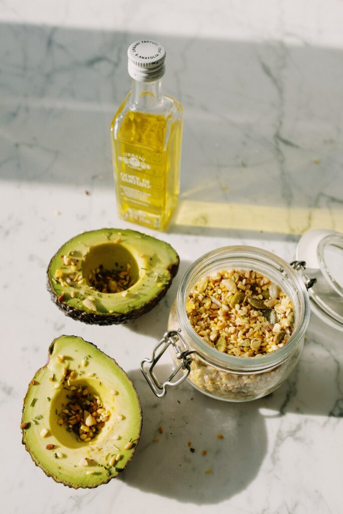 One Bottle of Olive Oil and a jar full of crushed nuts next to a Avocado cut in half with some of the nuts sprinkled over the AVO.